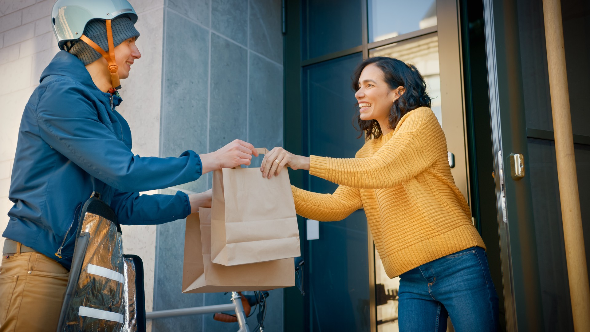 Happy Food Delivery Man on Bike Delivers Restaurant Order to Beautiful Female Customer. Courier Delivers Takeaway Lunch to Gorgeous Girl in Modern City District Office Building. Low Angle